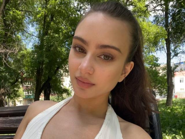 EleanorEvanchyk with tan skin, brown eyes, and dark brown hair in a ponytail, wearing a white halter top, smiling subtly, with small, round, nude-ass visible.