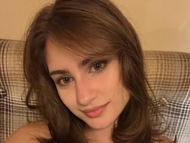 EleanorEvanchyk has brown hair, blue eyes, and a slight smile with pink lips. She's looking at the camera with her tits partially visible against a plaid backdrop.