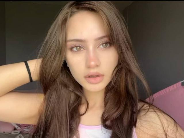 EleanorEvanchyk with long brown hair, green eyes, pink lips, wearing a pink tank top, black wristband, hand in hair, looking at camera with slightly parted pink lips.