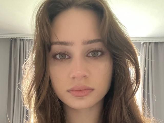 EleanorEvanchyk has wavy brown hair, green eyes, and pink lips. Her face is centered with a neutral expression. She has a light skin tone and subtle makeup.