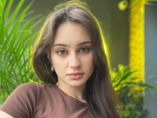 EleanorEvanchyk has brown hair, green eyes, and is wearing a brown shirt with earrings. She's staring directly at the camera with a neutral expression. Her tits are small.