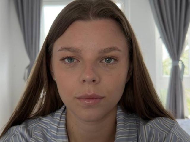 RhonaBells has pale skin, blue eyes, and long brown hair, wearing a striped shirt, with a neutral expression, in a bright room with gray curtains.