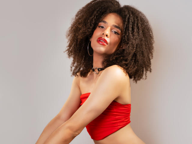 Kataleya Fowler met een grote, krullende afro, een rode top zonder bandjes, een zwarte choker, oorringen en rode lippenstift, leunt voorover, toont haar borsten en heeft een sexy, zelfverzekerde uitstraling..