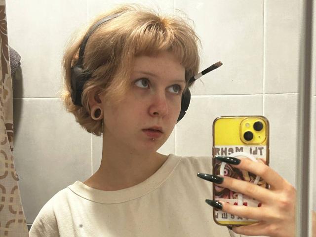 VasilikiAndersson with short blonde hair, wearing a headset, white shirt, and green nail polish, taking a mirror selfie with a yellow phone. Pierced nose, ear gauges.