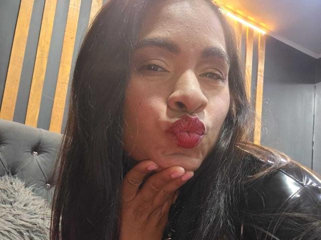 Emily Vidal, met lang zwart haar, die haar vingers kust, volle rode lippen, donkere wenkbrauwen, een zwarte leren jas, en bruine huid, houdt haar kin vast en kijkt naar de camera..