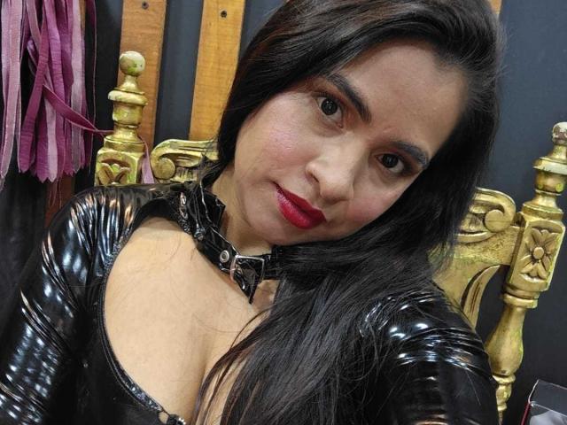 Emily Vidal, avec un haut en latex noir brillant, du rouge à lèvres rouge et des cheveux foncés, portant un collier en cuir noir, penchée en avant, montrant ses seins. Une chaise dorée et des sangles violettes sont visibles en arrière-plan..