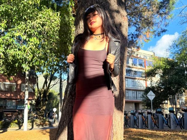 Mia Salim está de pie frente a un árbol, vestida con un vestido ajustado color burdeos, un collar negro y una chaqueta de cuero, con el pelo largo y negro y pintalabios rojo, con las manos sujetando la chaqueta..
