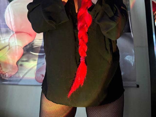AnnyTanny, con una coda di trecce rosso acceso, indossa una maglietta nera e calze a rete, ed è in piedi di fronte a un poster appeso al muro, che risulta sfocato. La sua maglietta è leggermente aperta..