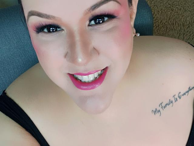 GiiannaMorrys with pink eyeshadow, bold pink lipstick, smiling, showing teeth, black bra, tattoo on right shoulder, short brown hair, small nose ring, fair skin, seated on grey couch.