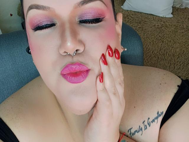 GiiannaMorrys with pink eyeshadow, red lipstick, nose ring, and red nails, pouting with hand on cheek, showing tattoo on left shoulder, sitting on grey chair.