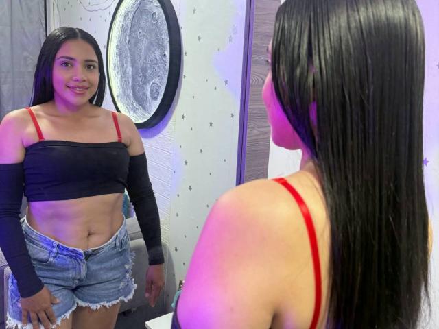 VickyRousse with long black hair, red bra, black off-shoulder top, and frayed denim shorts stands in front of a mirror, showing her bare belly and small tits.