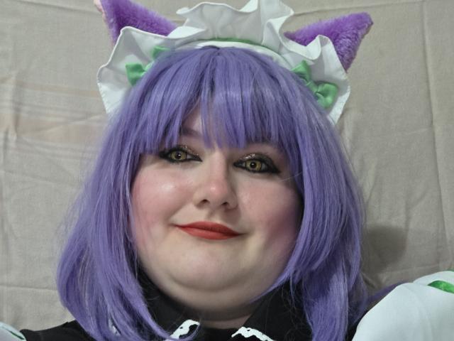 LaSexyChubby con orecchie da gatto viola, parrucca viola e copricapo da cameriera bianco, sorride con rossetto rosso, viso paffuto e trucco con occhi verdi, indossa un abito bianco e nero..