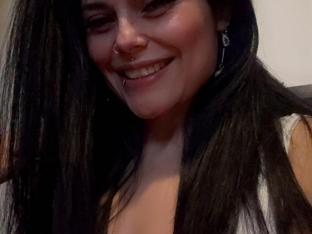 CharlotteCrofort with long black hair, smiling with a nose ring and lip piercing, showing her teeth, wearing a sleeveless top, and dangling earrings, looking cute and sexy.