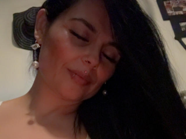 CharlotteCrofort with closed eyes, slight smile, black hair, pearl earrings, nose piercing, and a small lip piercing, wearing no clothes, in a dimly lit room.