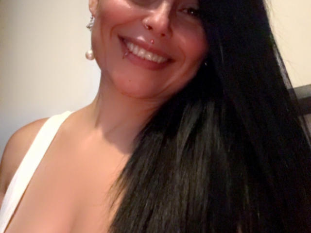 CharlotteCrofort with black hair, smiling, nose ring, pierced lip, white tank top, showing her tits, wearing pearl earrings, in a selfie with her face and upper body.