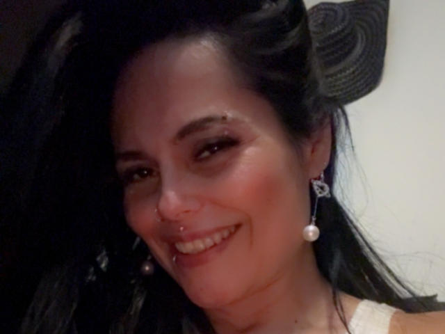 CharlotteCrofort has dark hair, a nose piercing, and a smiling face with pink blush, wearing pearl earrings and a white top, looking cute with a black hat.