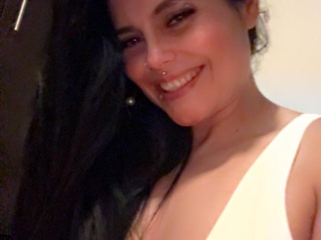 CharlotteCrofort with long black hair, smiling, nose ring, and white tank top, showing cleavage, has a pretty face, and is looking at the camera.