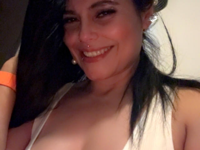 CharlotteCrofort with dark hair, smiling, nose ring, and earrings, wearing a white top, showing her tits, orange bracelet, and a slight blush on her face.