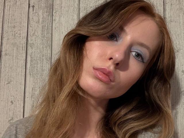 ReannaDreben has pouty pink lips, light blue eyeshadow, and wavy brown hair, leaning slightly to the side with a soft, relaxed expression. Her tits are not visible.