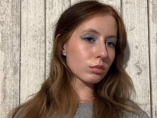 ReannaDreben a des fards à paupières bleus, des lèvres roses et gonflées, et de longs cheveux bruns. Elle porte une chemise grise et regarde légèrement sur le côté avec une expression neutre..