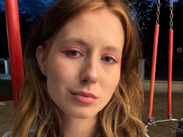 ReannaDreben ha la pelle chiara, l'eyeliner rosso e i capelli biondi ondulati, con un leggero sorriso, e si trova di fronte a delle altalene rosse al crepuscolo. Le sue labbra rosa sono leggermente dischiuse..