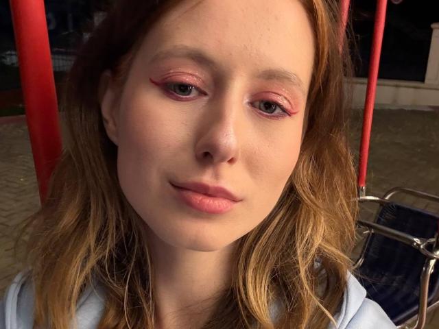 ReannaDreben's face, showing pink eyeliner, light brown wavy hair, pale skin, and pink lips, wearing a light blue hoodie, seated in front of red poles and a chair.
