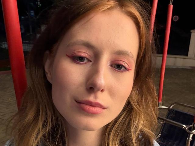 ReannaDreben with pale skin, red eyeliner, and pink eyeshadow, has wavy red hair and pink lips, looking slightly to the side with a subtle smile.