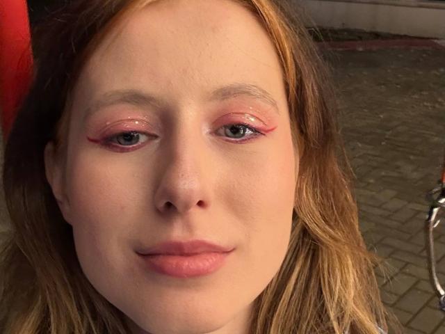 ReannaDreben's face, with pinkish eyeshadow, light brown wavy hair, and pink lips, showing a slight smile. Her nose is small, and her skin is fair.