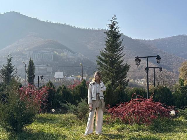 ReannaDreben stands in a grassy garden, wearing a white dress with a beige scarf, surrounded by green trees and red shrubs, with black lanterns in the background.