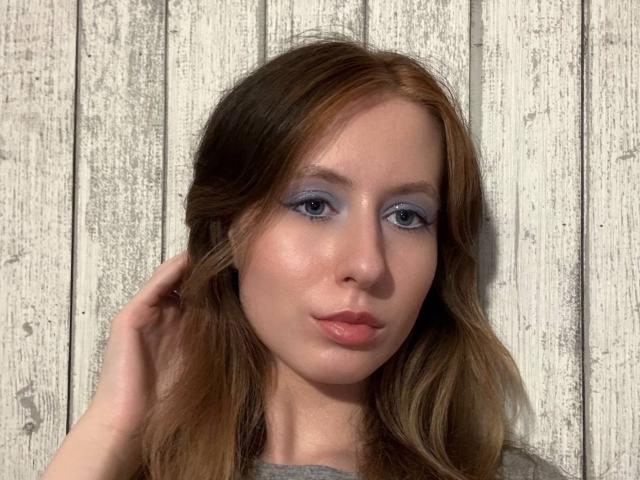 ReannaDreben's face, blue eyeshadow, pink lips, wavy brown hair, hand in hair, gray shirt, pale skin, looking at camera, wooden background.