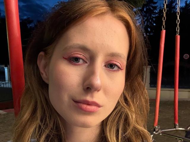 ReannaDreben with pink eyeshadow, light red lipstick, and wavy blonde hair, smiling slightly, standing in front of red swing bars at dusk. Her tits are small.