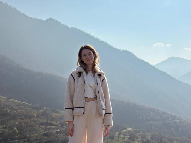 ReannaDreben stands in a mountain landscape wearing a fluffy white jacket with black trim, a white crop top, and high-waisted beige pants, looking relaxed with a slight smile.
