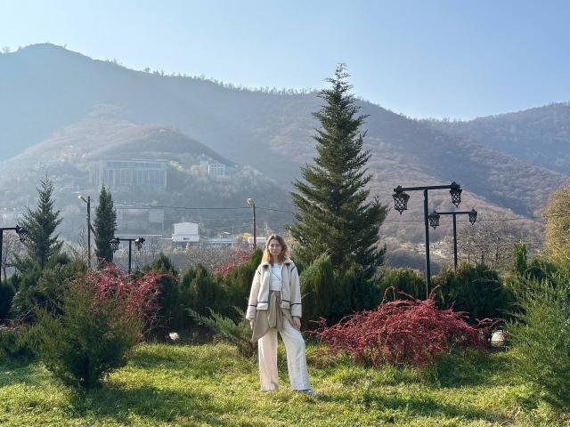 ReannaDreben stands in a grassy garden, wearing a beige coat and white pants, with red shrubs and green trees around, mountains and a clear sky in the background.