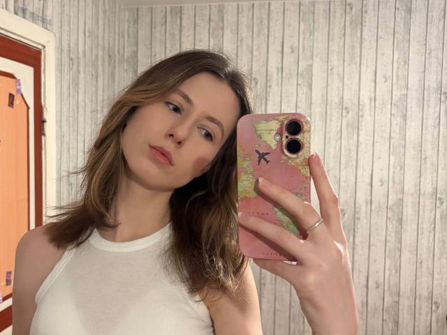 ReannaDreben, avec des cheveux bruns, un débardeur blanc, tenant un téléphone rose avec un motif de carte, prenant un selfie devant un miroir, montrant de petites seins, expression neutre, fond de mur en bois, alliance à la main gauche..