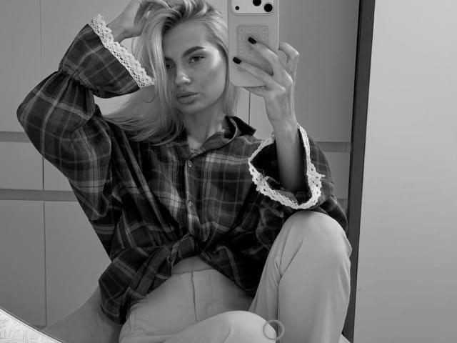 Pearlene Scalley, con i capelli biondi, indossa una camicia a quadri con polsini in pizzo, tiene in mano un telefono, sta scattando un selfie allo specchio, seduta con una gamba sollevata, in scala di grigi. Seni, sedere, vagina..
