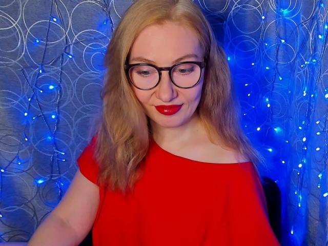 PaulinaSunny with red dress, black glasses, and red lipstick, has blonde hair, and is smiling with blue fairy lights in the background. Her tits are partially visible.