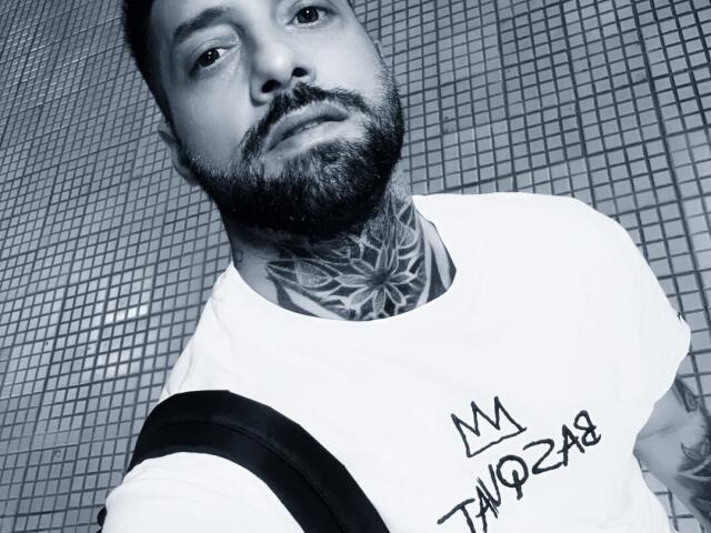 TommyDum tem uma barba grossa, tatuagens no pescoço e uma camisa branca com uma coroa e "TQ258" em preto. Ele está usando uma alça preta sobre o ombro..