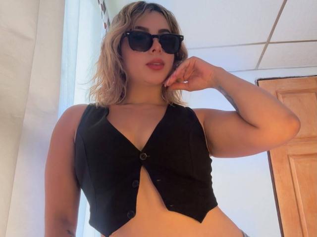 CloeEvanss with wavy blonde hair, black sunglasses, and a black sleeveless vest showing her tits, standing with her right hand near her face.