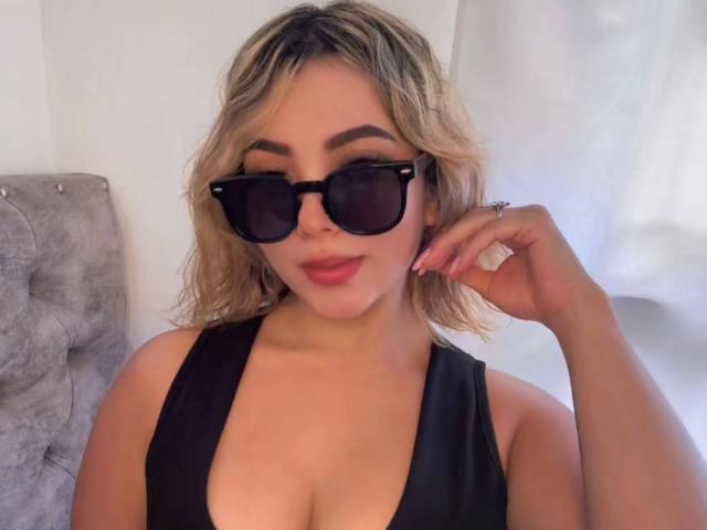 CloeEvanss with blonde wavy hair, wearing black sunglasses, black low-cut top, showing her boobs, holding her right hand up, pink lipstick, and a ring on her finger.