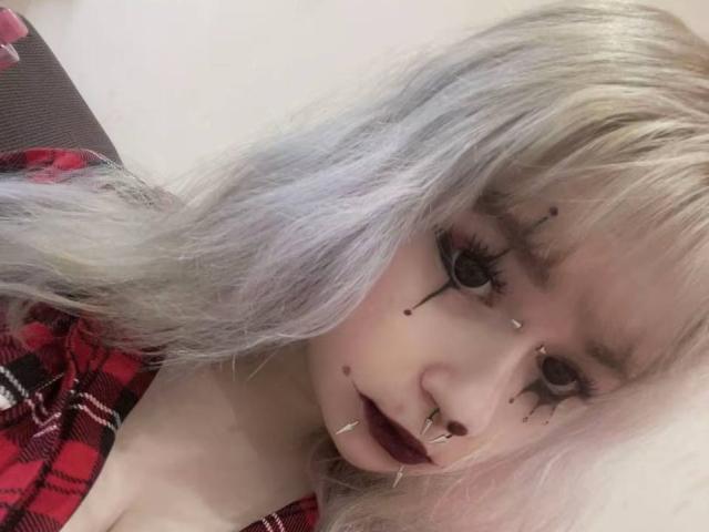 EmmaKoven with platinum blonde hair, goth makeup, black eyeliner, nose piercings, dark lipstick, and red plaid shirt. Her face is adorned with black dots and small metallic pieces.