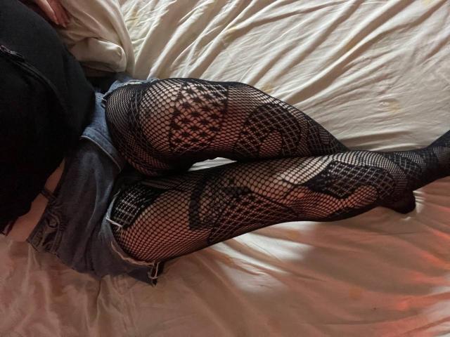 EmmaKoven's legs in black fishnet stockings with floral patterns, lying on a white bed. She's wearing denim shorts, black top, and her ass is partially visible.
