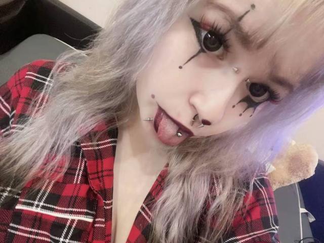 EmmaKoven with pale skin, purple-gray hair, black eyeliner, and dark lipstick, sticking out tongue with nose and lip piercings, wearing red plaid shirt, in selfie.