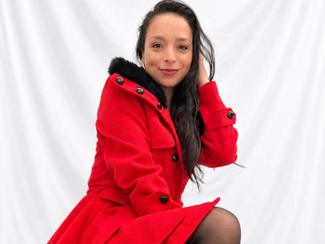 StangelarRivera with long dark hair, smiling, wearing a red coat with black fur collar, black buttons, and black stockings, sitting, hand in hair.