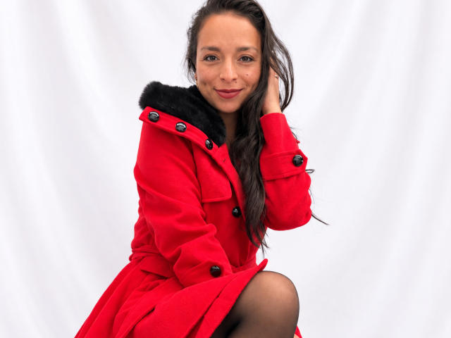 StangelarRivera has long brown hair, a cute smile, and is wearing a red coat with black fur trim, squatting with one hand in her hair, showing her black panty.