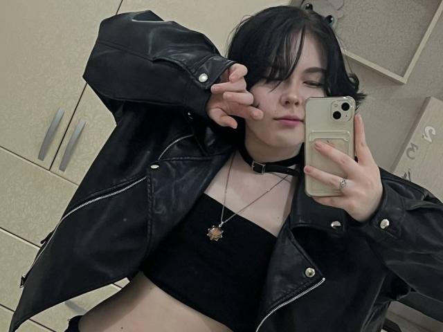 EvitaCorolis, with black leather jacket, black choker, black strap top, and short black hair, takes a mirror selfie, showing off her tits and a pendant, in a room.