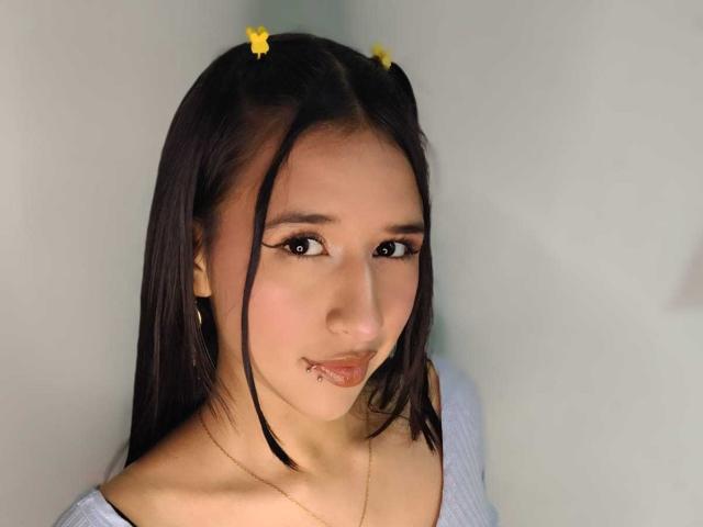 LilyRossi has straight black hair with yellow butterfly clips, a gold necklace, and a lip piercing. She's wearing a white top, looking cute with eyeliner and subtle makeup.
