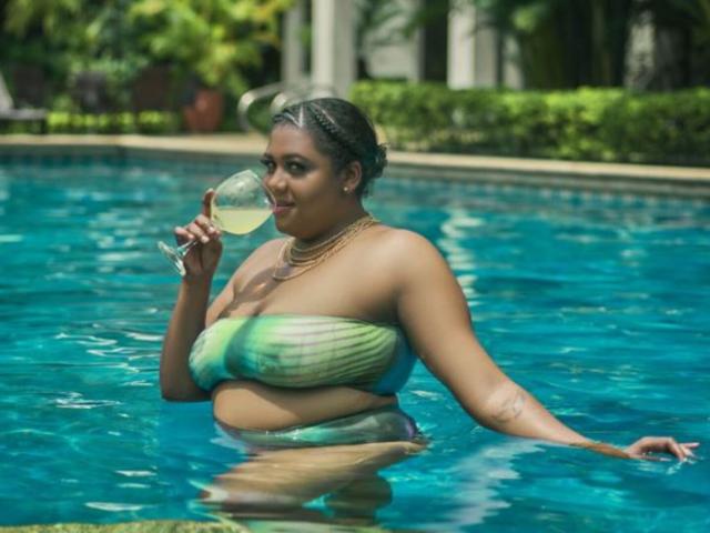 BiancaForest with big ass and big tits, wearing a green and blue striped bikini, holding a glass of white wine, standing in a pool, smiling.