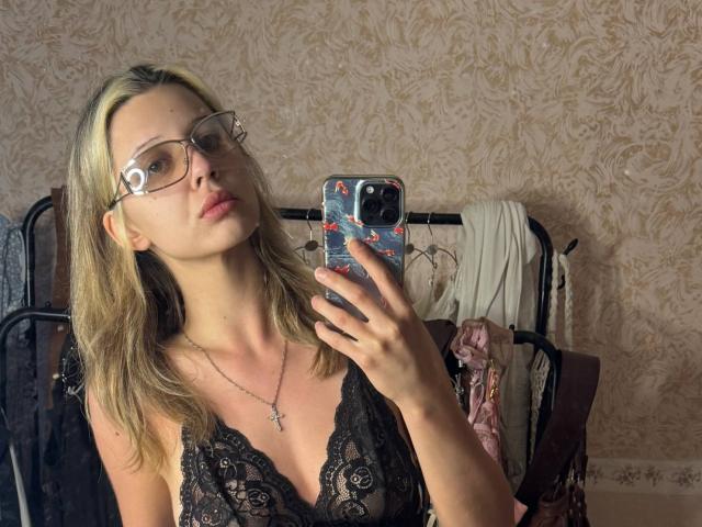 MerrieHamad, com cabelo loiro, óculos e blusa preta de renda, segurando um telefone, mostrando os seios, usando um colar com uma cruz, selfie no espelho, quarto com papel de parede estampado, roupas ao fundo..