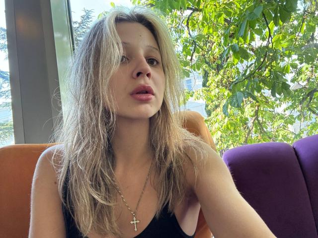 MerrieHamad, con el cabello rubio despeinado, un top negro, un collar con una cruz y los labios ligeramente entreabiertos, sentada en un compartimento iluminado por el sol, de color naranja y púrpura, con hojas verdes afuera..