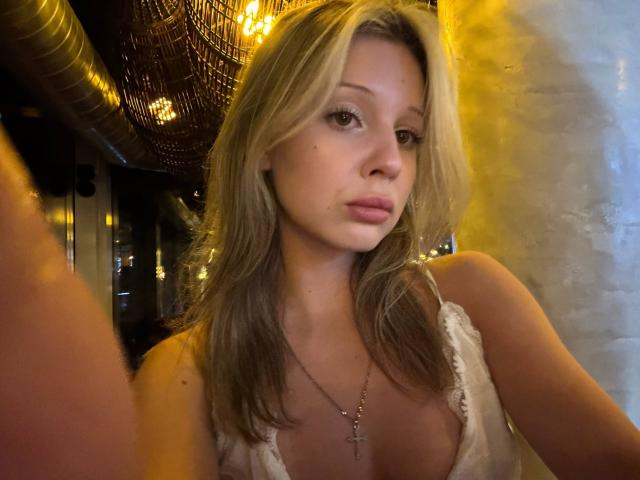 MerrieHamad with blonde hair, small tits, wearing a white top, cross necklace, looking at camera, yellow-lit background, metal duct, and hanging light visible.